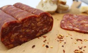 En bit spianata calabrese med synliga bitar av chili och nötter, bredvid en skiva och lite bröd.