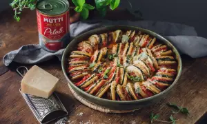 En vacker ugnsbakad ratatouille, en klassisk vegetarisk italiensk mat, med skivade grönsaker och ost. Bredvid står en burk Mutti tomater, en bit parmesan och en rivare.