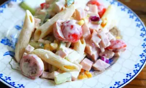 En krämig pastasallad med skinka, penne, körsbärstomater, gurka och majs på en tallrik med blått mönster.