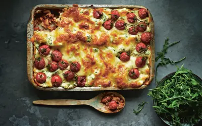 En gyllene salsiccia lasagne toppad med körsbärstomater och ost. En sked med lasagne och en skål rucola bredvid.
