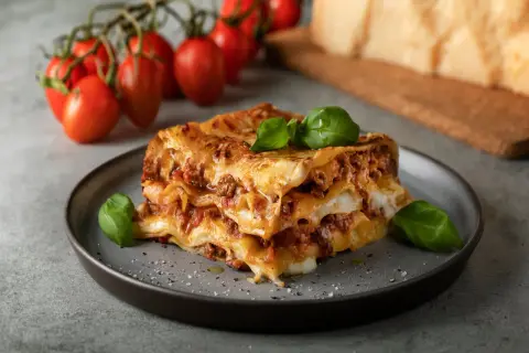 En bit italiensk lasagne med köttfärssås och ost, garnerad med basilika. Tomater och parmesanost i bakgrunden.