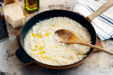 Krämig italiensk risotto i en panna, med en träsked som rör om. Parmesan och olivolja syns i bakgrunden.