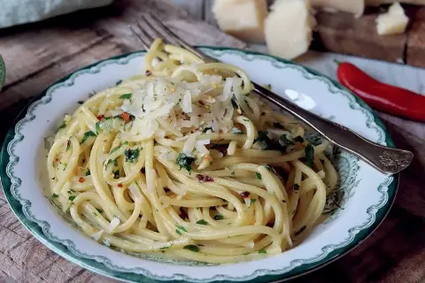 En tallrik med spaghetti aglio e olio, toppad med riven parmesan och persilja. En röd chili och ostbitar syns i bakgrunden.