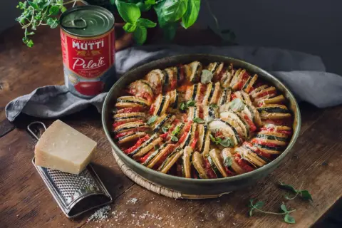 En vacker ugnsbakad ratatouille, en klassisk vegetarisk italiensk mat, med skivade grönsaker och ost. Bredvid står en burk Mutti tomater, en bit parmesan och en rivare.