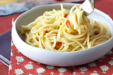 En skål med spaghetti aglio olio e peperoncino, där en gaffel lyfter upp en del av den oljiga pastan med små röda chilistrimlor.