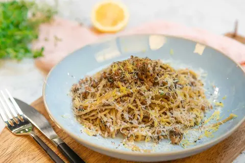 En tallrik med saffranspasta vegetarisk, toppad med riven ost och citronzest. Serveras med bestick på en träskärbräda.