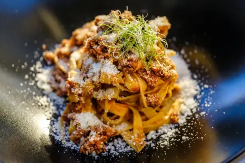 En portion pappardelle al cinghiale, toppad med riven ost och färska groddar, serverad på en svart tallrik.