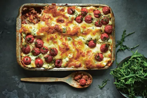 En gyllene salsiccia lasagne toppad med körsbärstomater och ost. En sked med lasagne och en skål rucola bredvid.