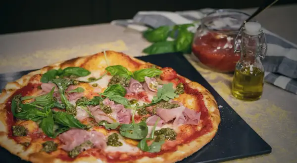 En läcker spenatpizza med prosciutto, basilika och pesto. Bredvid står en burk tomatsås och en flaska olivolja.