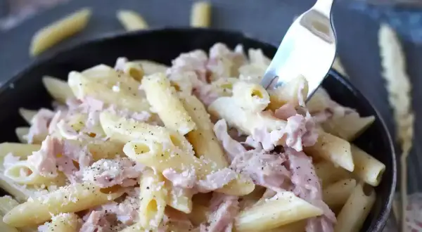En gaffel lyfter en tugga av krämig skinksås pasta, toppad med riven ost och svartpeppar.