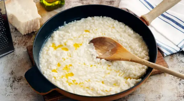Krämig italiensk risotto i en panna, med en träsked som rör om. Parmesan och olivolja syns i bakgrunden.