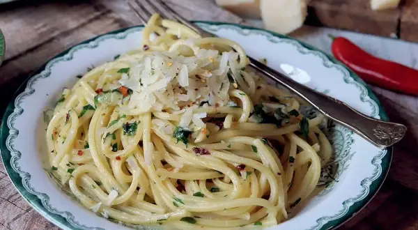 En tallrik med spaghetti aglio e olio, toppad med riven parmesan och persilja. En röd chili och ostbitar syns i bakgrunden.