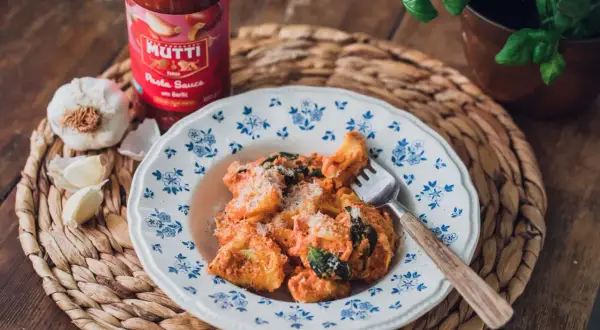 En tallrik med pasta i krämig tomatsås, toppad med parmesan. Bredvid står en burk Mutti pastasås och färsk vitlök. En perfekt italiensk meny fest!