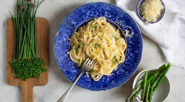 En tallrik krämig pasta limone, toppad med riven ost och persilja. Bredvid finns färsk gräslök och sparris.
