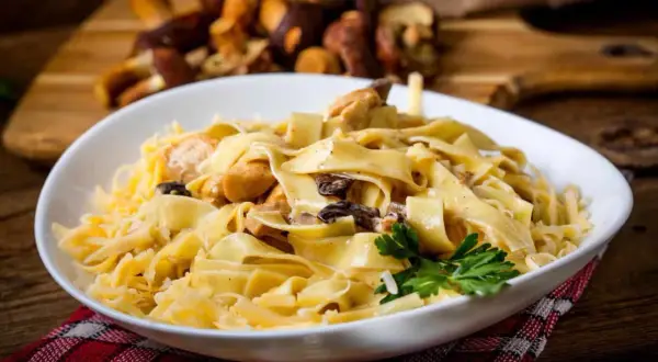 En skål med krämig pasta med kyckling och svamp, toppad med riven ost och en persiljekvist. I bakgrunden syns färska karljohanssvampar. En härlig pasta porcini-rätt.