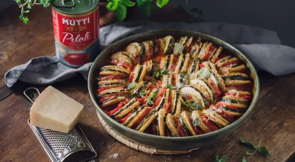 En vacker ugnsbakad ratatouille, en klassisk vegetarisk italiensk mat, med skivade grönsaker och ost. Bredvid står en burk Mutti tomater, en bit parmesan och en rivare.