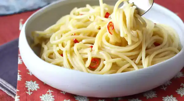 En skål med spaghetti aglio olio e peperoncino, där en gaffel lyfter upp en del av den oljiga pastan med små röda chilistrimlor.