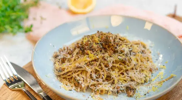 En tallrik med saffranspasta vegetarisk, toppad med riven ost och citronzest. Serveras med bestick på en träskärbräda.