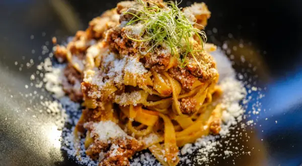 En portion pappardelle al cinghiale, toppad med riven ost och färska groddar, serverad på en svart tallrik.