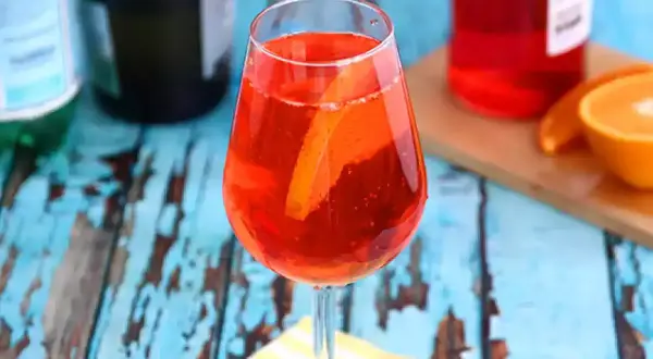En Aperol Spritz, Italiens nationaldryck, med apelsinskiva i glaset. Bakom syns flaskor och en apelsinhalva.