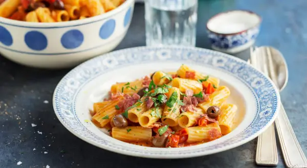 En tallrik med rigatoni pasta med salami, oliver och tomater, toppad med färsk persilja. En skål med mer pasta syns i bakgrunden.