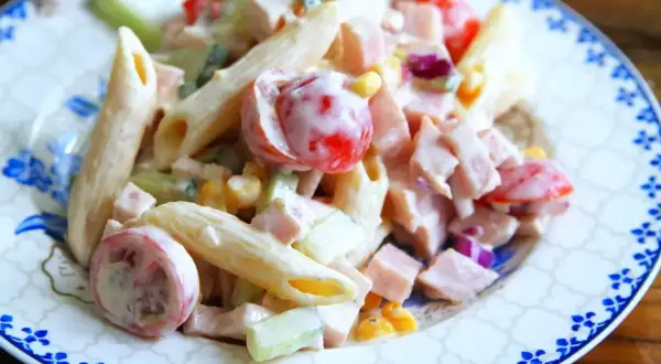 En krämig pastasallad med skinka, penne, körsbärstomater, gurka och majs på en tallrik med blått mönster.