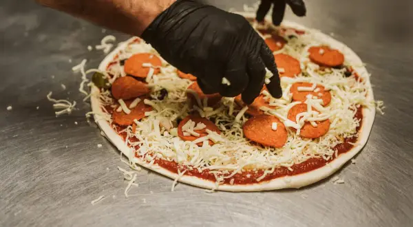 Händer i svarta handskar lägger ost på en pizza med pepperoni och oliver. En klassisk kombination för härliga pizza smaker.