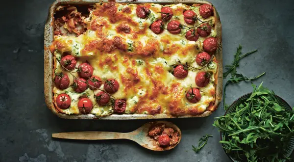 En gyllene salsiccia lasagne toppad med körsbärstomater och ost. En sked med lasagne och en skål rucola bredvid.