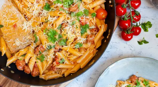 En stor panna med krämig pasta med chorizo, toppad med riven ost och färsk persilja. Bredvid syns en tallrik med samma läckra pasta.