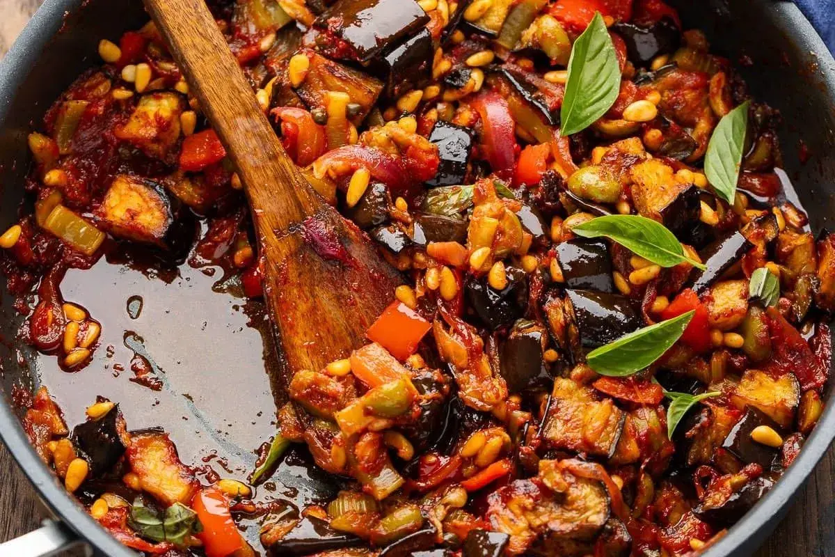 En varm, mustig siciliansk caponata med aubergine, paprika och pinjenötter i en panna, redo att serveras.