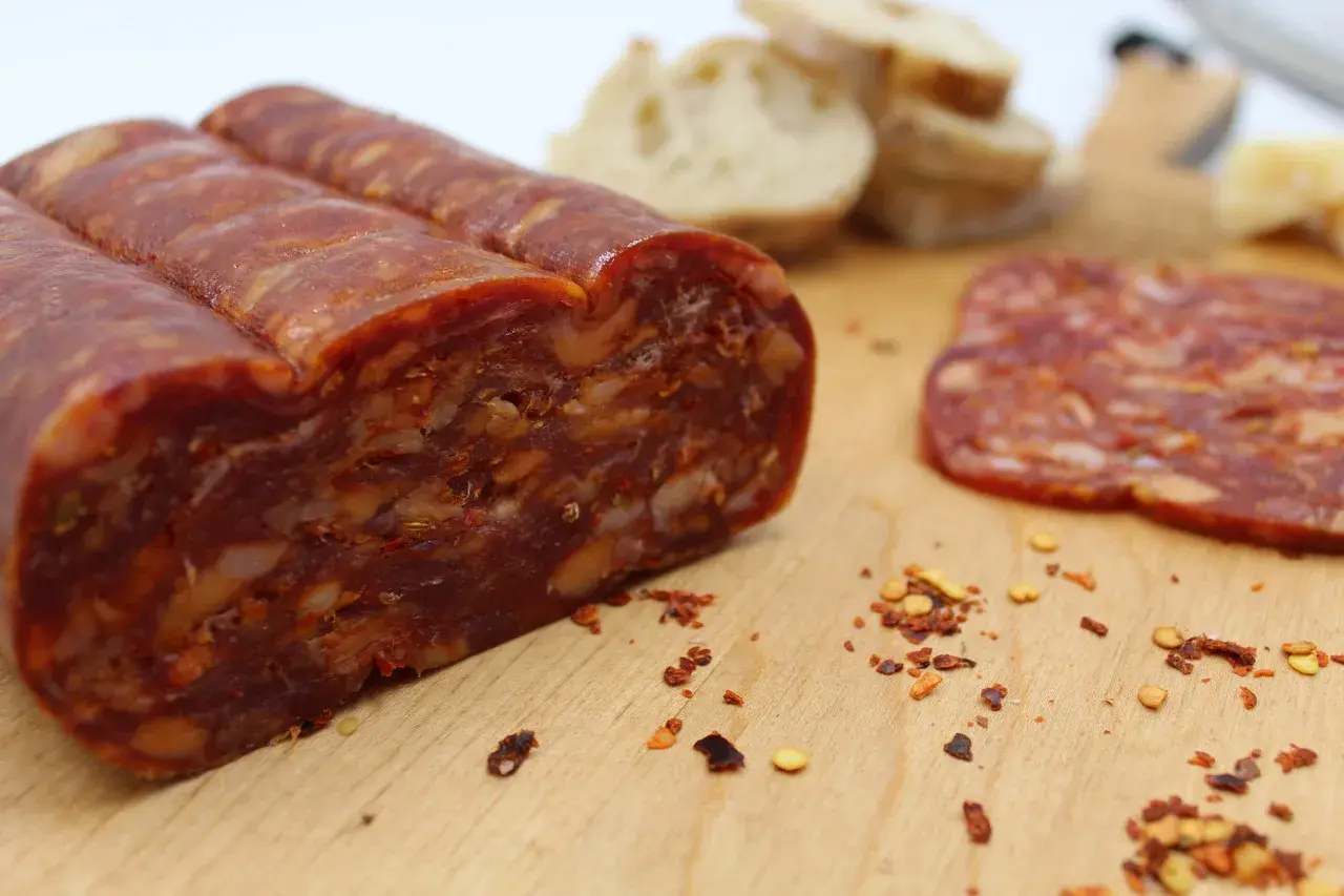 En bit spianata calabrese med synliga bitar av chili och nötter, bredvid en skiva och lite bröd.