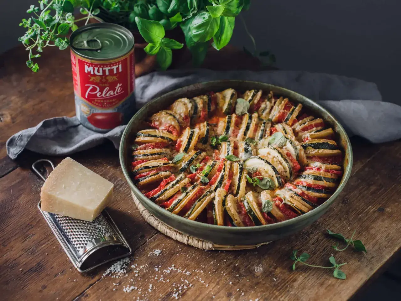 En vacker ugnsbakad ratatouille, en klassisk vegetarisk italiensk mat, med skivade grönsaker och ost. Bredvid står en burk Mutti tomater, en bit parmesan och en rivare.
