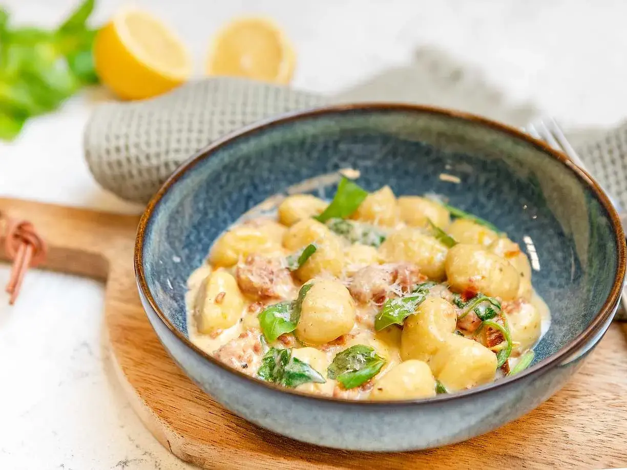 En skål med krämig gnocchi salsiccia, toppad med färsk basilika och parmesan. Citroner och en grå handduk i bakgrunden.