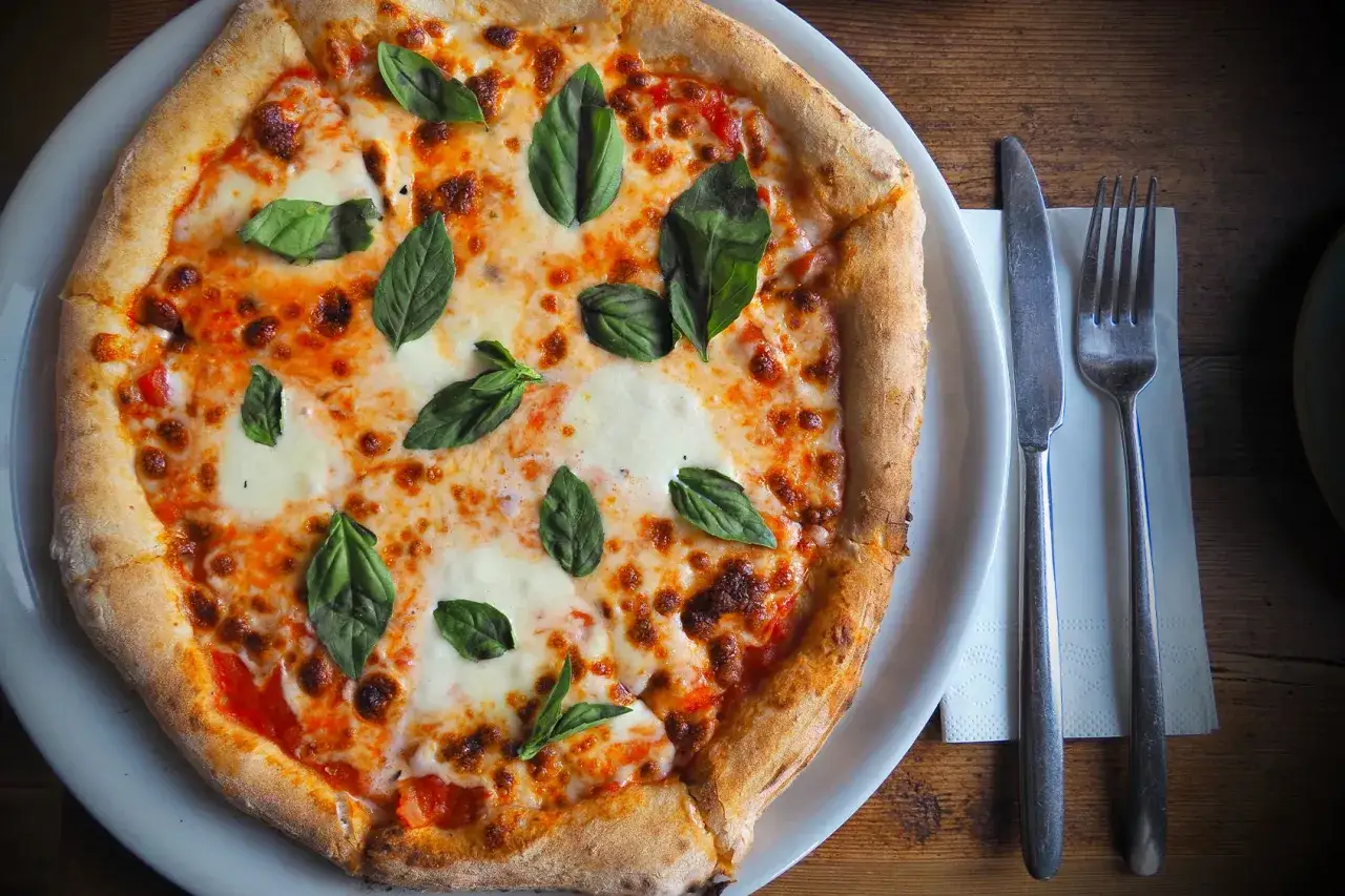 En pizza med tomatsås, rikligt med smält mozzarella liknande ost och färsk basilika. Bredvid ligger en kniv och gaffel.