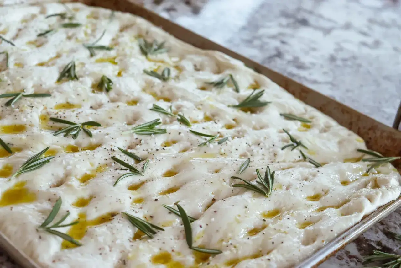 Focaccia rosmarin på en plåt, redo att bakas. Olivolja, flingsalt och peppar toppar det fluffiga brödet.