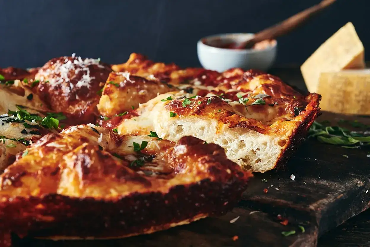 En gyllene, bubblande pizza i stekpanna med smält ost och färska örter. En bit är uppskuren och visar den luftiga bottnen.