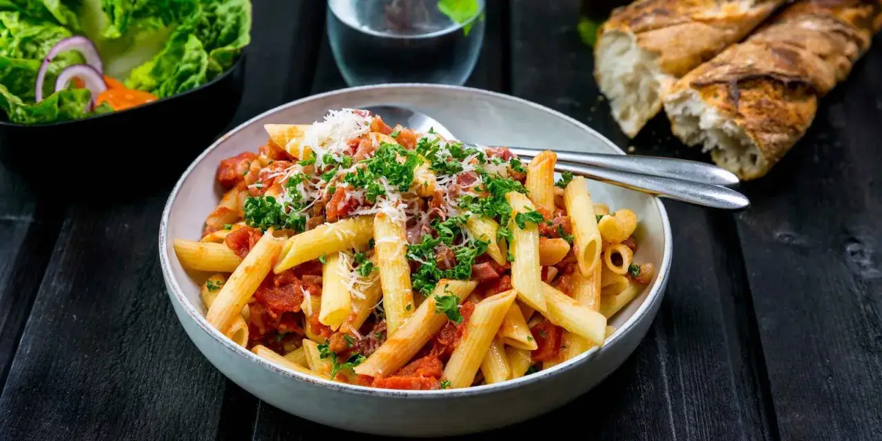 En skål med penne all'arrabiata, toppad med persilja och parmesan. Bredvid en sallad och ett glas vatten.