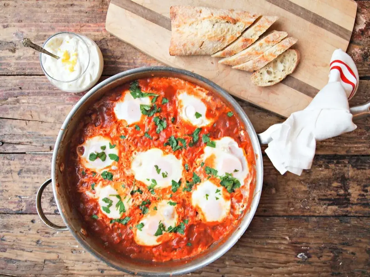 En smaklig shakshuka med ägg i tomatsås, serverad med bröd och en klick gräddfil. Ett perfekt shakshuka recept för helgfrukosten.