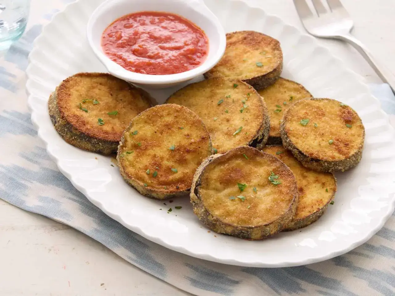 Krispigt fritera aubergine på en vit tallrik med en skål tomatsås. Garnerad med persilja.