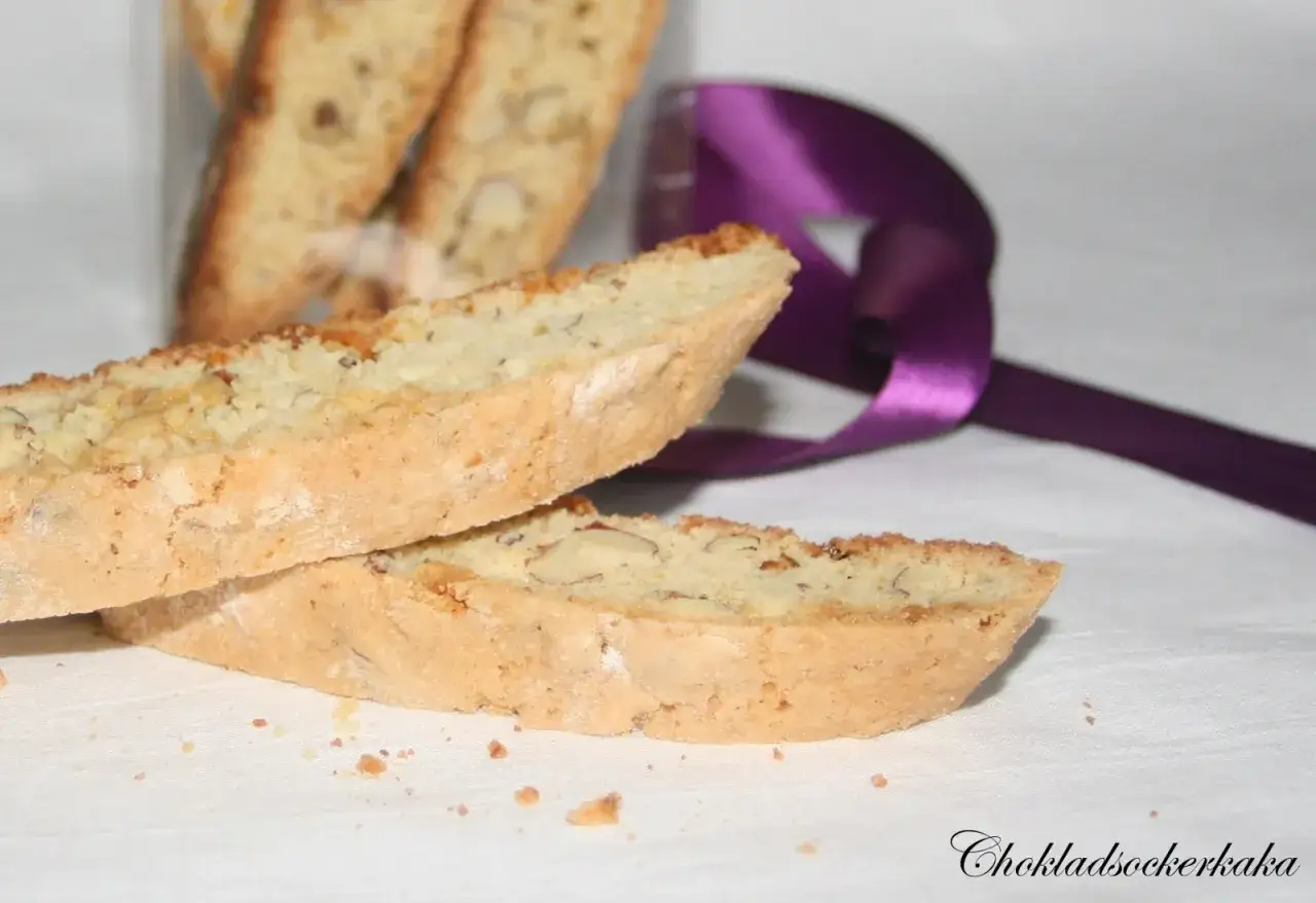 Två mandel-biscottis ligger på en vit duk, med en lila sidenband i bakgrunden. Ett perfekt recept på biscotti!