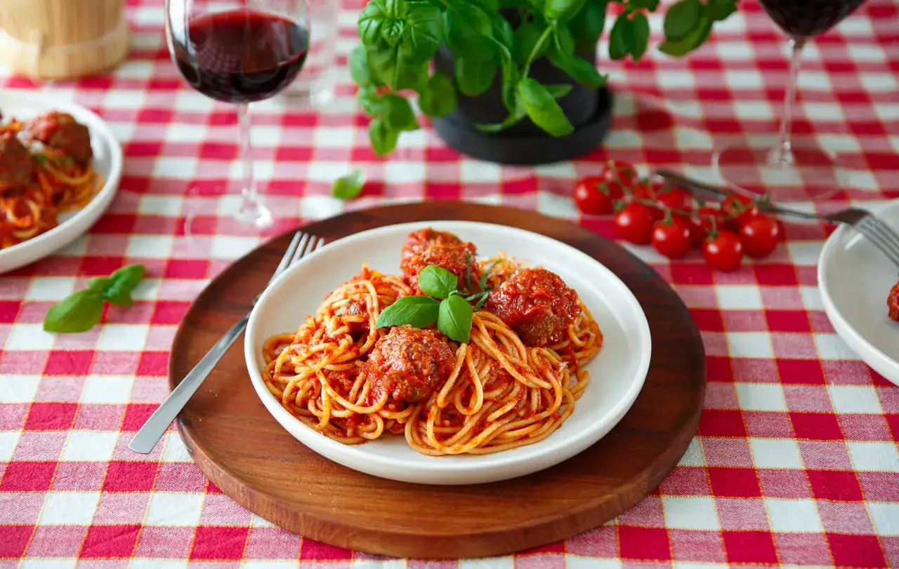 En tallrik med spaghetti och köttbullar (polpette) i tomatsås, garnerad med basilika. Röd rutig duk och ett glas rödvin i bakgrunden.