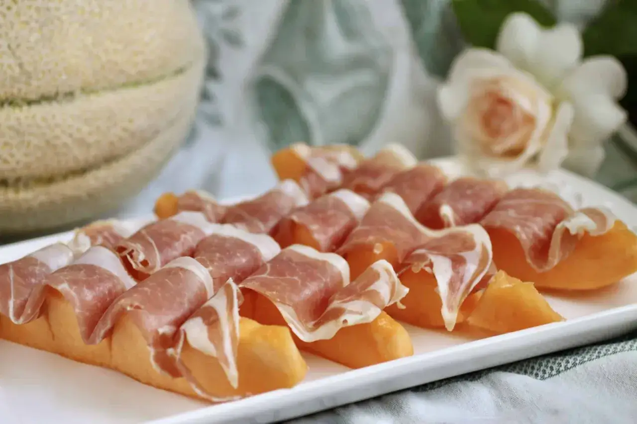Melonbitar inslagna i tunt skivad prosciutto på en vit tallrik. En hel melon syns i bakgrunden.