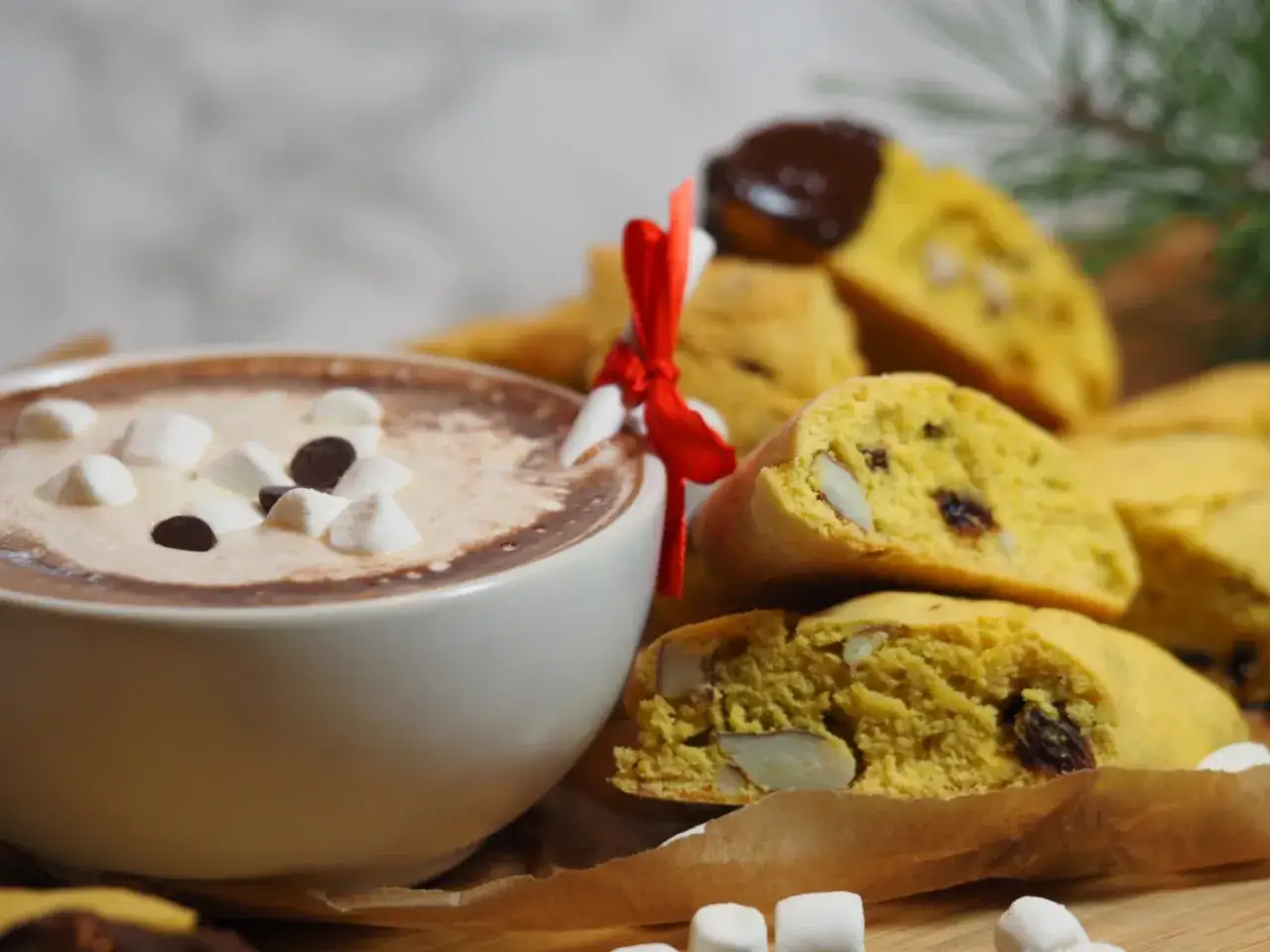 En kopp varm choklad med marshmallows och chokladbitar bredvid en hög med saffransbiscotti med mandel, bundna med ett rött band.