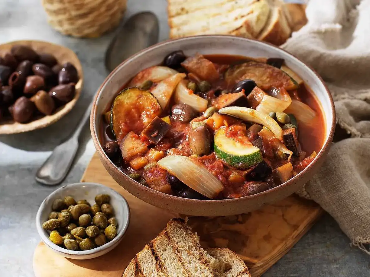 En skål med siciliansk caponata, full av aubergine, zucchini, oliver och kapris. Serveras med rostat bröd.
