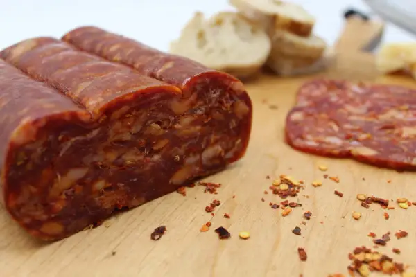 En bit spianata calabrese med synliga bitar av chili och nötter, bredvid en skiva och lite bröd.