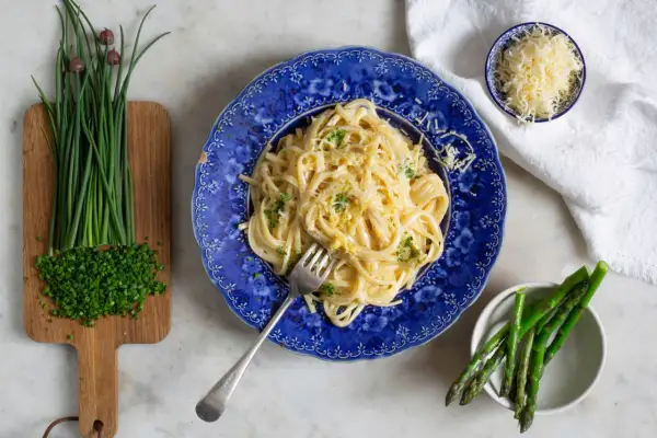 En tallrik krämig pasta limone, toppad med riven ost och persilja. Bredvid finns färsk gräslök och sparris.