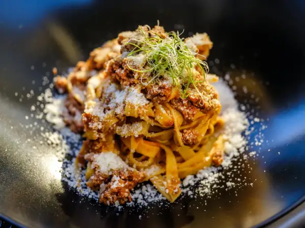 En portion pappardelle al cinghiale, toppad med riven ost och färska groddar, serverad på en svart tallrik.