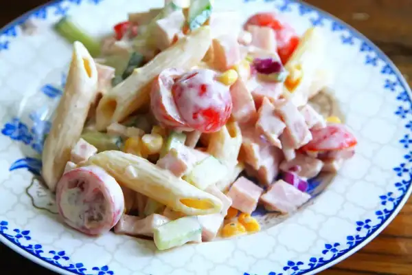 En krämig pastasallad med skinka, penne, körsbärstomater, gurka och majs på en tallrik med blått mönster.
